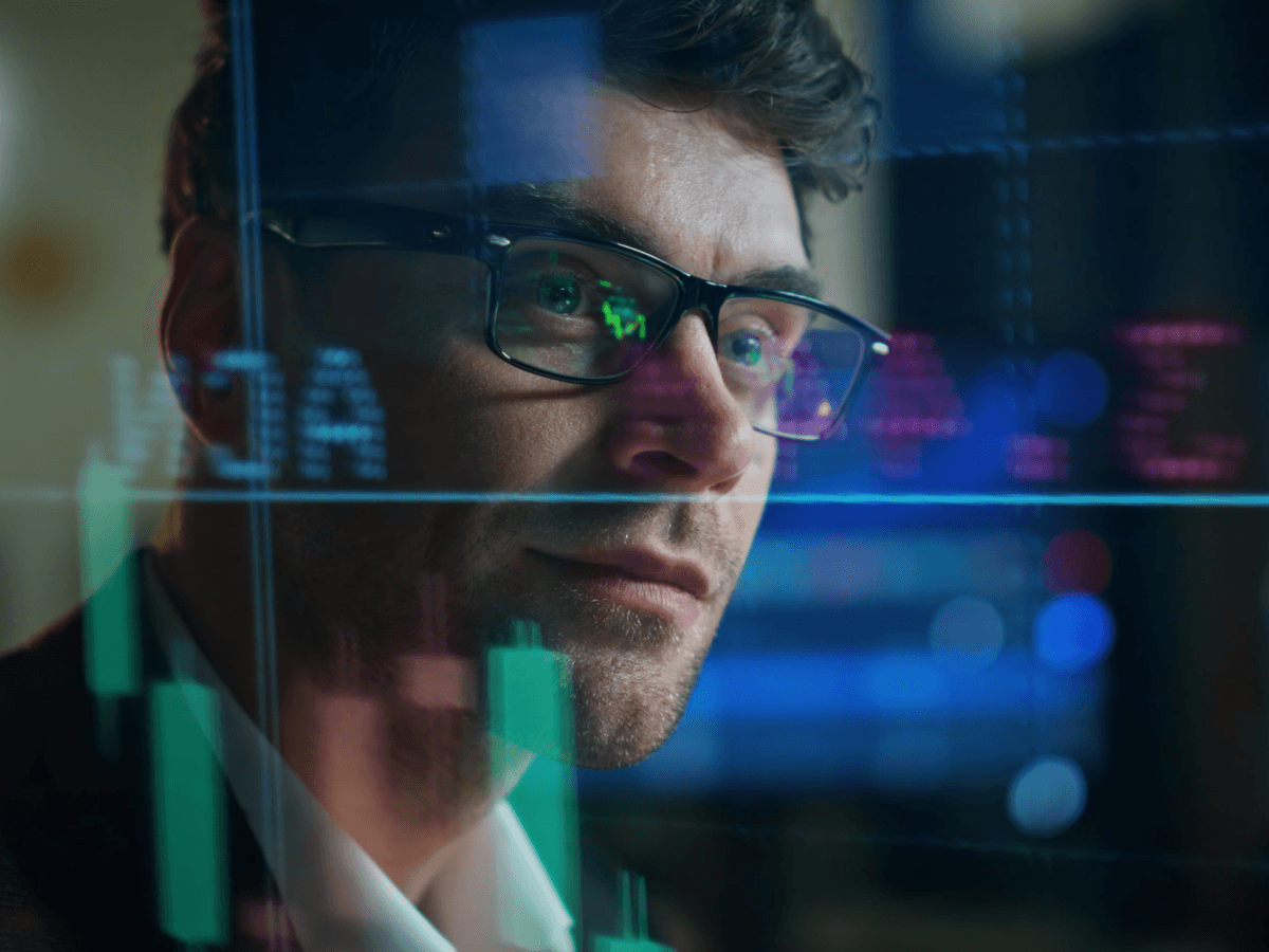A man wearing glasses analyzes stock market data on a computer screen, focused on the information presented.